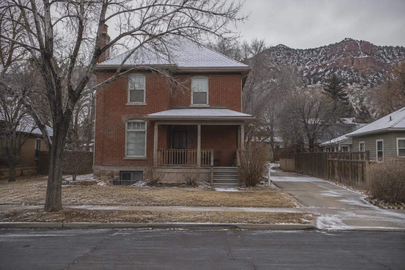 Older red brick home under bare trees with a light snow in Cedar City, Utah